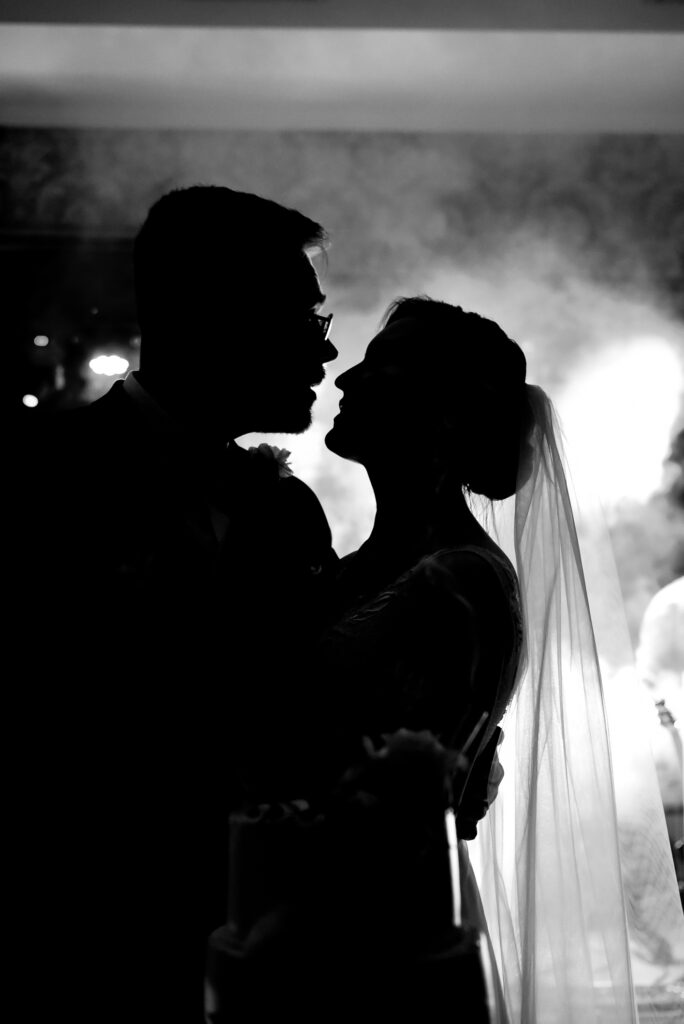 Czarnobiały obraz przedstawia sylwetki dwóch osób, prawdopodobnie pary, stojących blisko siebie. Osoba po prawej stronie ma na głowie welon, co sugeruje scenę ślubną. Tło zdjęcia jest wypełnione dymem lub mgłą, co tworzy dramatyczny efekt, a źródło światła za postaciami dodatkowo podkreśla ich zarys.