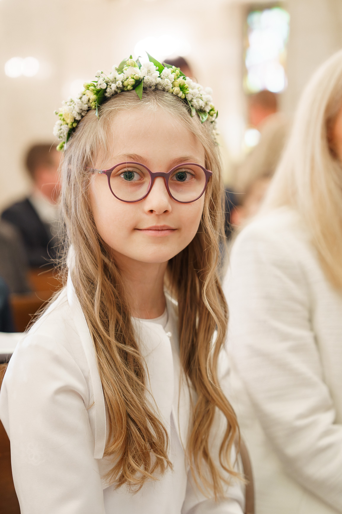 Dziewczynka z długimi blond włosami, ubrana w białą sukienkę komunijną, z ozdobnym wiankiem z białych kwiatów i zielonych liści na głowie. Tło rozmyte, prawdopodobnie przedstawia wnętrze kościoła lub salę podczas ceremonii religijnej, z innymi osobami w oddali.