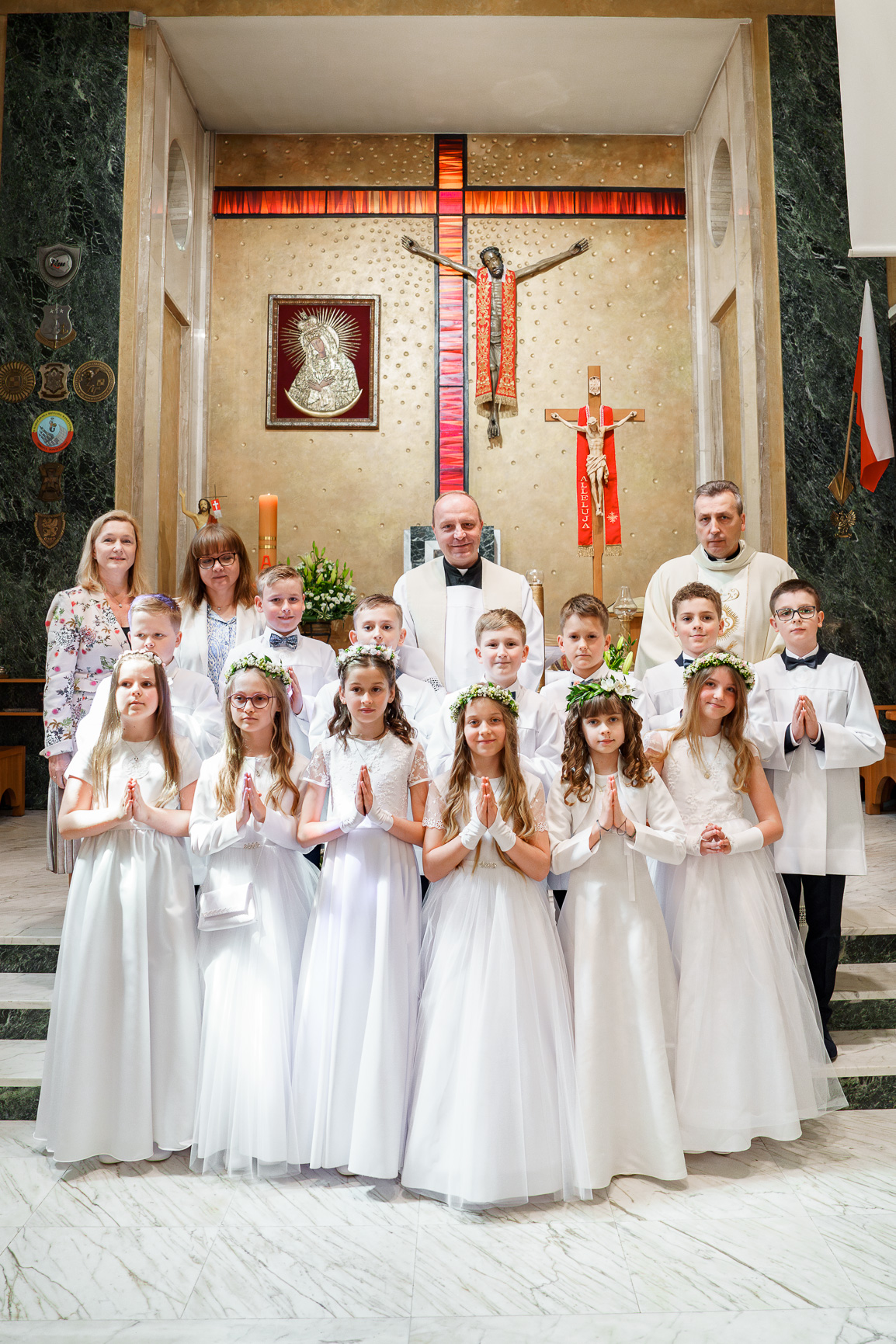 Grupa dzieci w białych albach ustawiona w półokręgu przed ołtarzem w kościele podczas uroczystości Pierwszej Komunii Świętej. W tle widać dekoracje kwiatowe, zapalone świeczki oraz krzyż na ścianie.